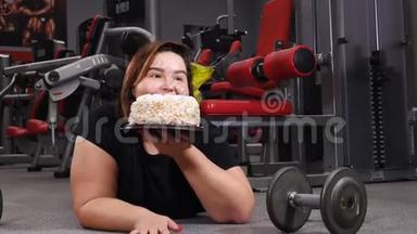 有趣的肥胖女人在健身房拿着美味的蛋糕。 穿着黑色t恤的胖女人躺在空健身房的地板上，上面有一个哑铃